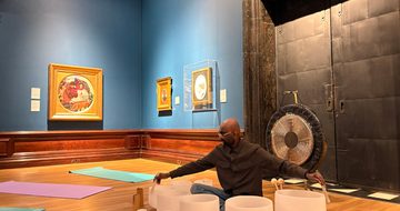 Man seated on wooden floor playing white crystal singing bowls with mallets, surrounded by Pre-Raphaelite paintings.