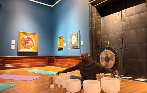 Man seated on wooden floor playing white crystal singing bowls with mallets, surrounded by Pre-Raphaelite paintings.