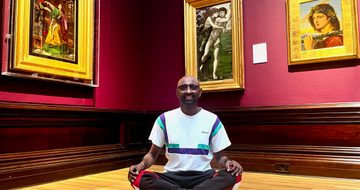 A person in a meditation yoga pose in an art gallery - paintings in gold frames are on the walls behind.