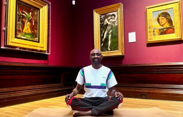 A person in a meditation yoga pose in an art gallery - paintings in gold frames are on the walls behind.