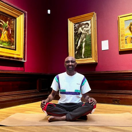 A person in a meditation yoga pose in an art gallery - paintings in gold frames are on the walls behind.