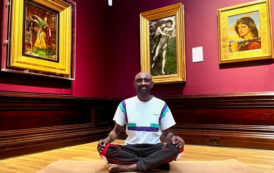 A person in a meditation yoga pose in an art gallery - paintings in gold frames are on the walls behind.
