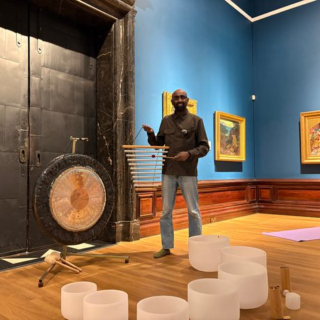 Man standing beside a gong and crystal singing bowls in an art gallery room.