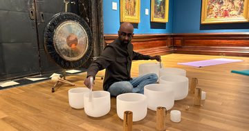 Man seated on wooden floor playing singing bowls with mallets, surrounded by Pre-Raphaelite paintings.