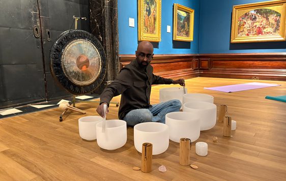 Man seated on wooden floor playing singing bowls with mallets, surrounded by Pre-Raphaelite paintings.