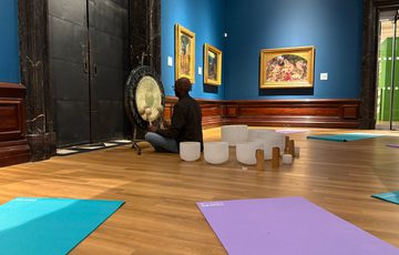 Person plays a gong in an art gallery, surrounded by singing bowls, yoga mats and Pre-Raphaelite paintings on the walls.