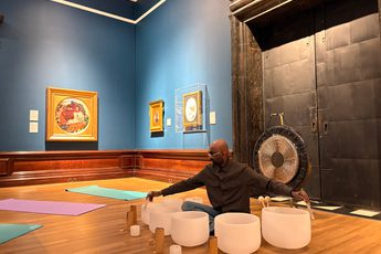 Man seated on wooden floor playing white crystal singing bowls with mallets, surrounded by Pre-Raphaelite paintings.