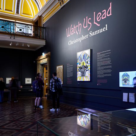 A wide shot of a gallery display with people looking at the large stained glass artwork.