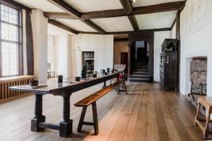 Historic Tudor building with wooden floors, large wooden table in centre, fireplace, window and staircase.