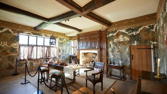 Inside a historic room with wooden beams and floor, a fire place and table anc chairs.