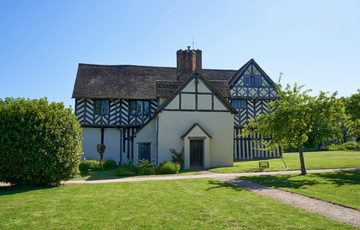Exterior side view of a Tudor timber-framed house and garden with lawn and trees.