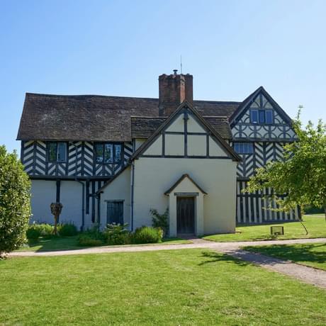 Exterior side view of a Tudor timber-framed house and garden with lawn and trees.