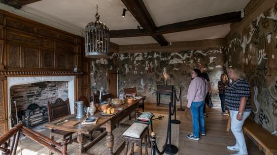 Visitors in a Tudor dining room looking at the dining table surrounded by chairs and stools in front of unlit fireplace.