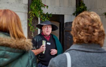Close up of the back of the heads of two visitors who are listening to a tour guide who is dressed in a hat and cape.