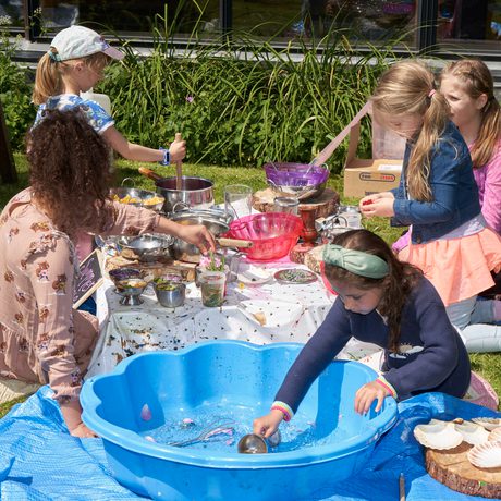 A group of children sitting on the grass with a low table in front of them and plastic sandpit with water. The children are playing with bowls and bits of plants and making things.