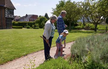 Two adults and a child are looking at plants in a garden. The child holds a clipboard. Blakesley Hall Tudor building is background.