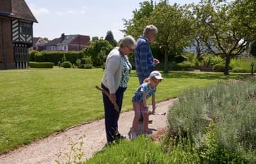 Two adults and a child are looking at plants in a garden. The child holds a clipboard. Blakesley Hall Tudor building is background.
