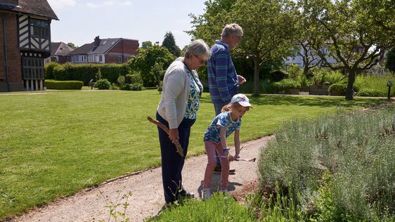 Two adults and a child are looking at plants in a garden. The child holds a clipboard. Blakesley Hall Tudor building is background.