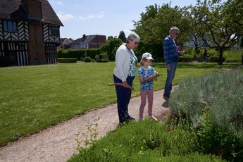 Two adults and a child are looking at plants in a garden. The child holds a clipboard. Blakesley Hall Tudor building is background.