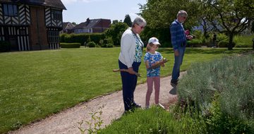 Two adults and a child are looking at plants in a garden. The child holds a clipboard. Blakesley Hall Tudor building is background.