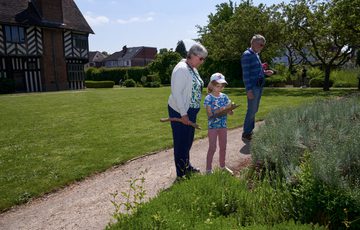 Two adults and a child are looking at plants in a garden. The child holds a clipboard. Blakesley Hall Tudor building is background.