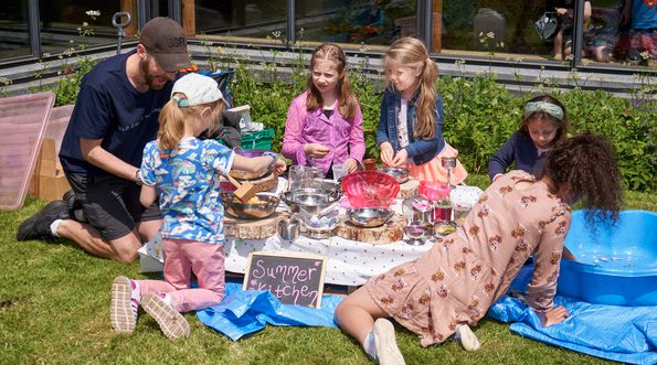A group of children and a adult are outside. A sign reads 'summer kitchen'. The children are playing with bowls and saucepans and natural items such as petals and foliage.