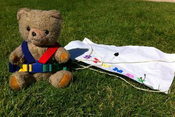 A teddy wearing a harness and paper parachute sitting on the grass.