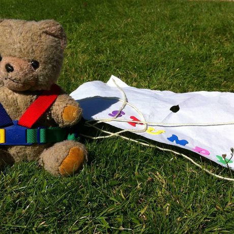 A teddy wearing a harness and paper parachute sitting on the grass.
