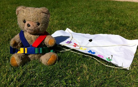 A teddy wearing a harness and paper parachute sitting on the grass.