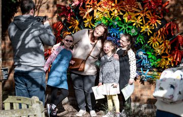 An adult and three children pose for a photo taken by another adult. They are standing in front of a flowered rainbow artwork.