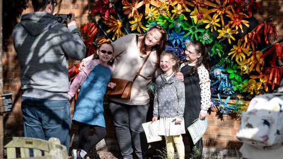 An adult and three children pose for a photo taken by another adult. They are standing in front of a flowered rainbow artwork.
