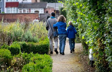 A family inlcuding a adult and two children, walking down a path surrounded by shrubs. They face away from the camera.