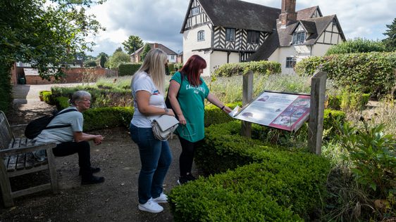 Two ladies reading an information in the garden. Another person sits on a nearby bench. Blakesley Hall Tudor building is in the background.