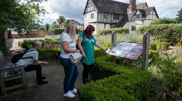 Two ladies reading an information in the garden. Another person sits on a nearby bench. Blakesley Hall Tudor building is in the background.