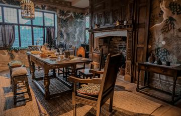 Inside a historic room with wooden beams and floor, a fire place and table and chairs and large window.