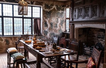 Inside a historic room with wooden beams and floor, a fire place and table and chairs and large window.