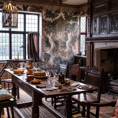Inside a historic room with wooden beams and floor, a fire place and table and chairs and large window.