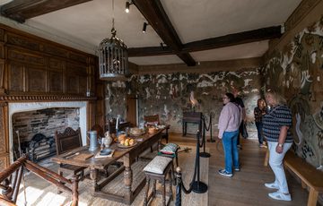 Visitors in a Tudor dining room looking at the dining table surrounded by chairs and stools in front of unlit fireplace.