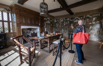 A lady looking at the ceiling of a Tudor dining room. In the room is a table surrounded by chairs and stools in front of unlit fireplace.