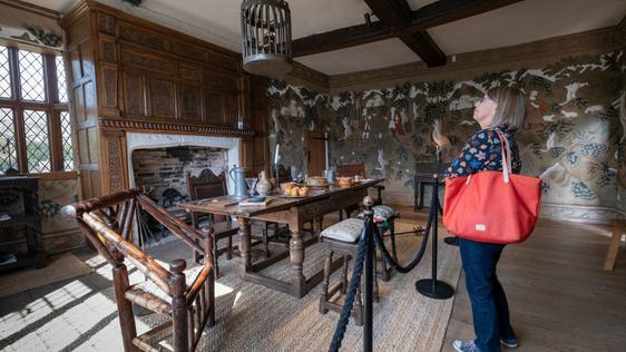 A lady looking at the ceiling of a Tudor dining room. In the room is a table surrounded by chairs and stools in front of unlit fireplace.