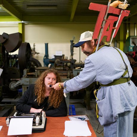 A man in blue outfit with a "TAT" sign on his back, offers a microphone to a person seated behind a typewriter.