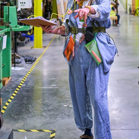 Man in eclectic blue outfit gestures while holding clipboard in industrial museum store.