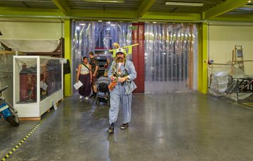 Person in blue outfit and 'TAT' sign on his back, walks in a large, industrial museum store space with following behind.
