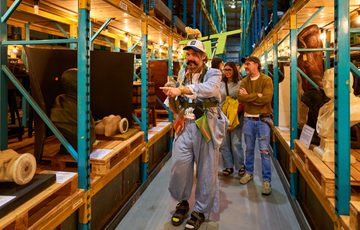 Man in blue outfit and "TAT" sign on his back, leads group through museum aisle filled with artefacts.