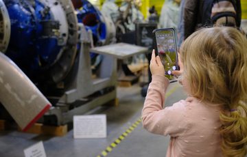 A child holding a phone and taking a photo of a museum object or engine that is on display.