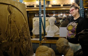 A perpson smiling while looking at a a selection of museum objects including busts that are on display in the warehouse on shelves.