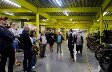 A person is giving a tour to a group of people in a warehouse with vintage vehicles to the side.