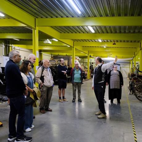 A person is giving a tour to a group of people in a warehouse with vintage vehicles to the side.