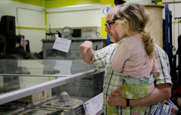 An adult holding a child so they can get a better look at display of miniature buildings that are in a perspex case.