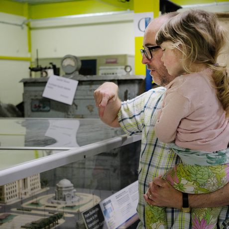 An adult holding a child so they can get a better look at display of miniature buildings that are in a perspex case.